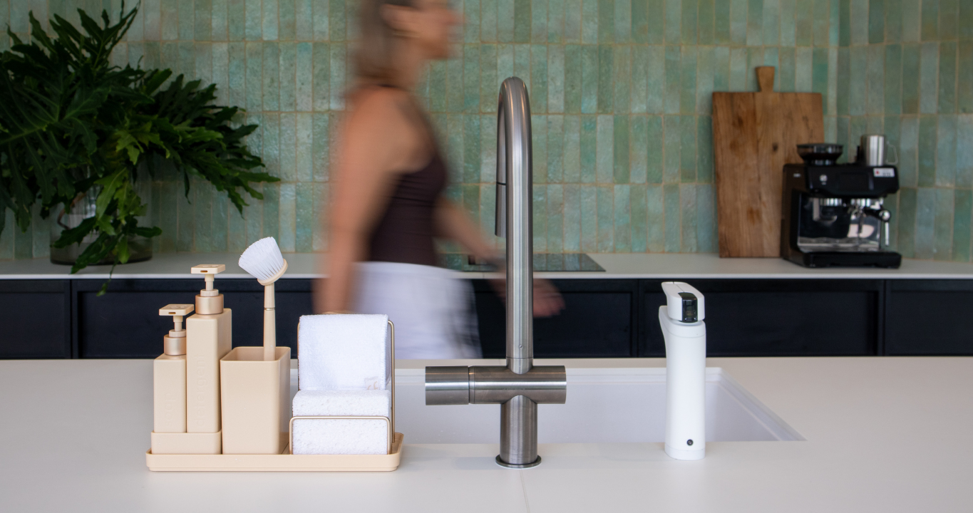 Modern kitchen sink with faucet and cleaning products on a countertop, blurred person in the background.